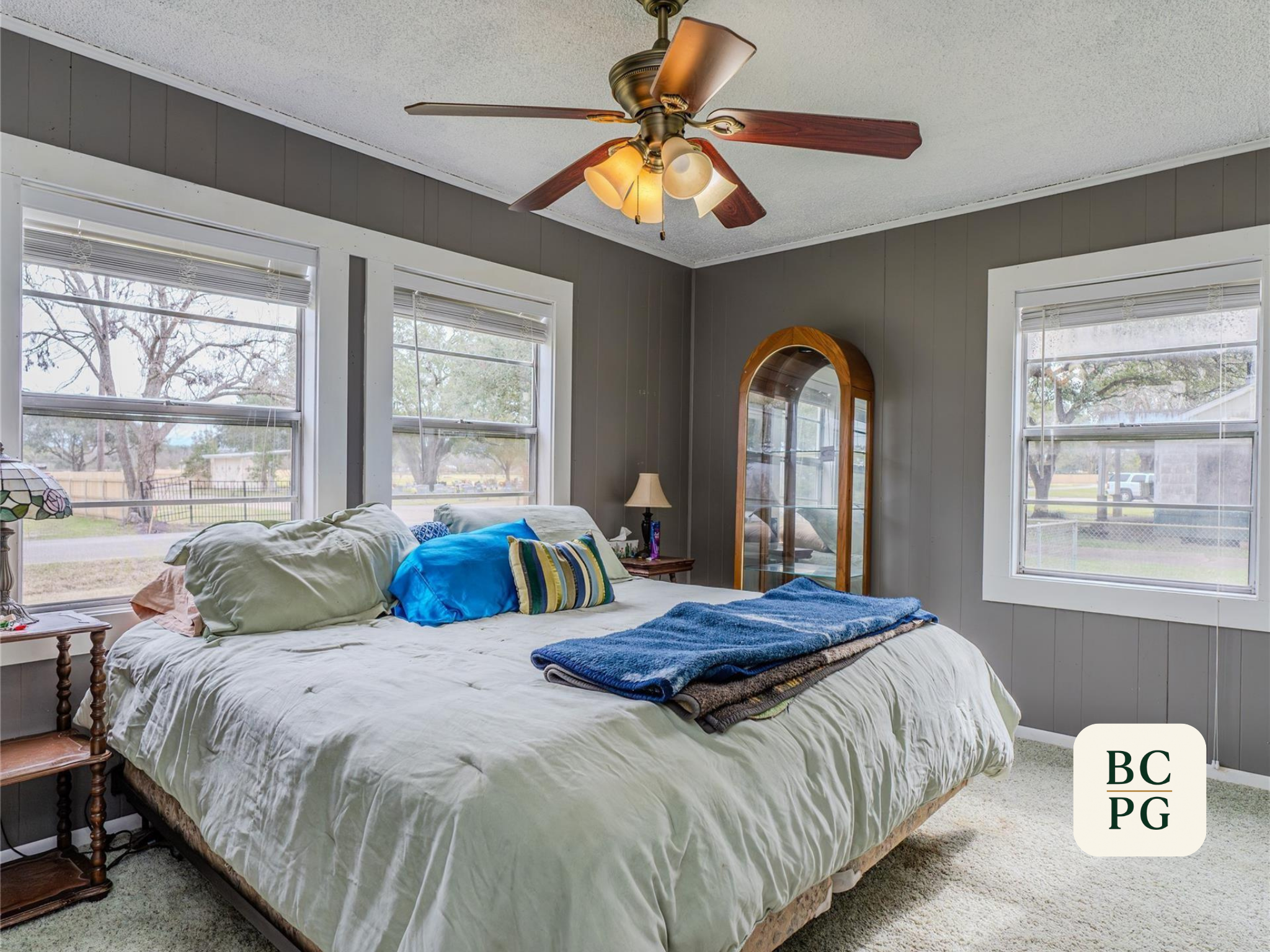 A bedroom with a bed that has white sheets, various pillows, and a folded blanket on top. There is a wooden side table with a lamp next to the bed. The room features three large windows with white blinds, letting in natural light. A ceiling fan with 