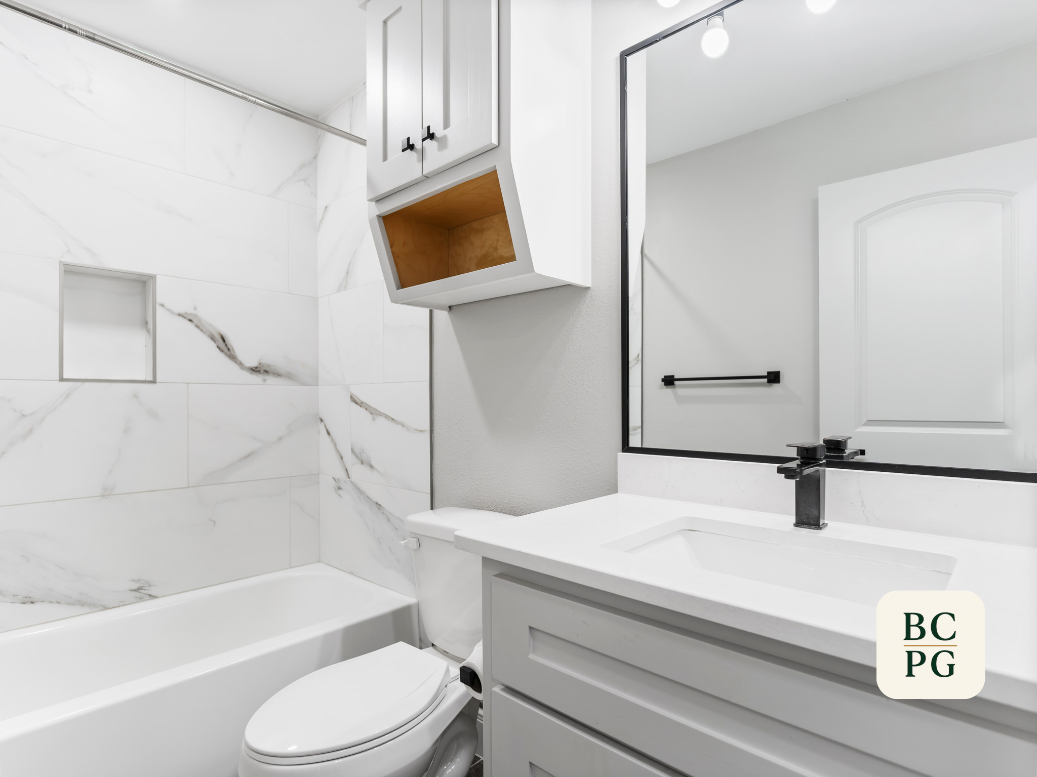 A modern bathroom with white marble walls, a white bathtub, a white vanity with a black faucet, a large mirror, black towel rack, and wall-mounted storage cabinet with open shelves.