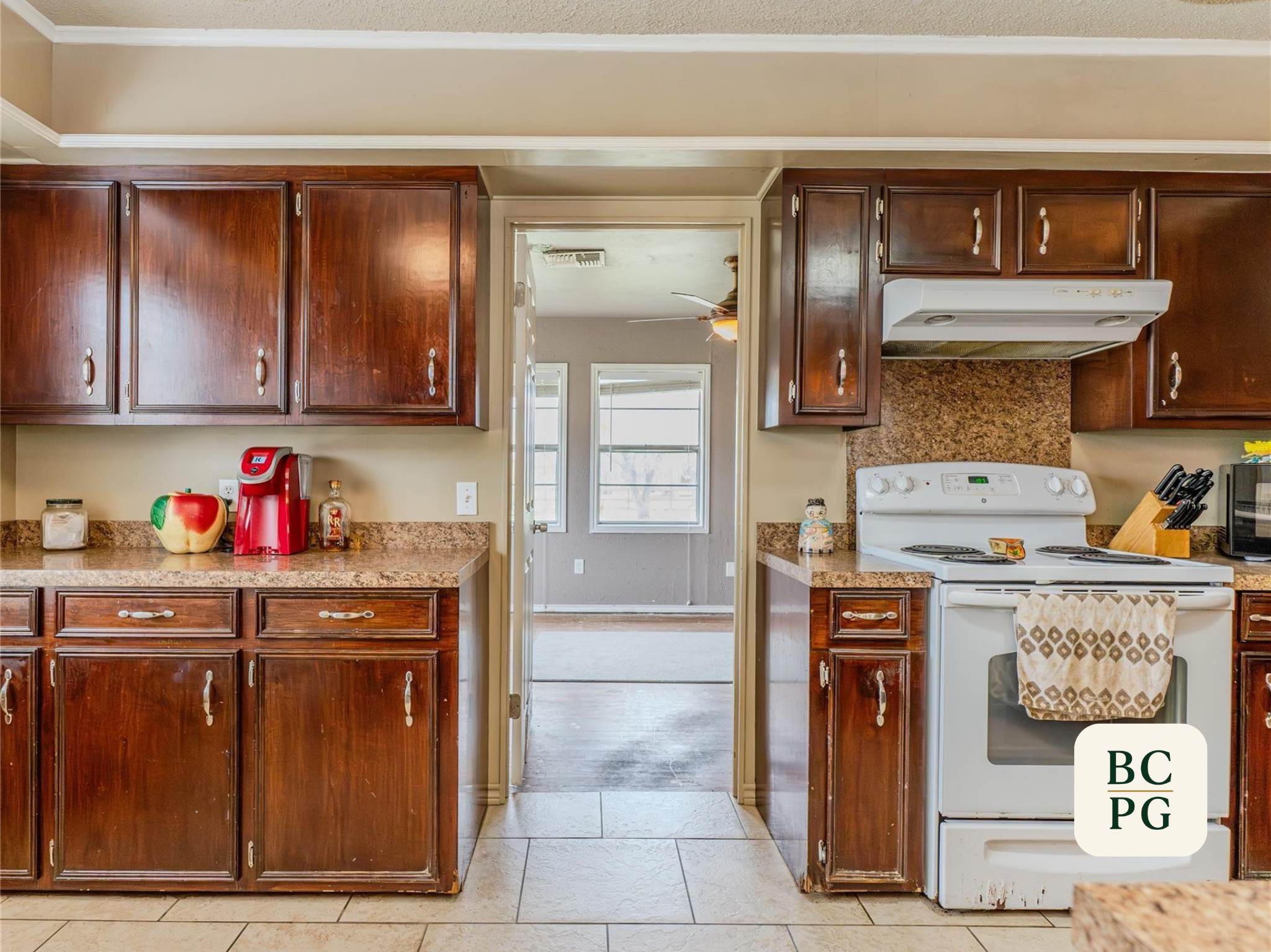 View of a kitchen with wooden cabinets, a stove, and a doorway leading to another room with a person holding a long object. The countertop has a red coffee maker, an apple-shaped container, a small liquor bottle, and a decorative figurine. There are 