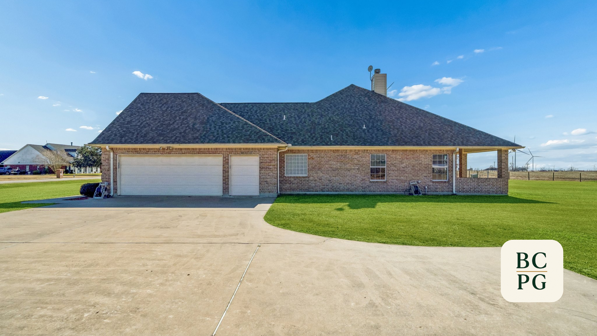 A brick house with a dark shingled roof, white garage doors, large windows, and a chimney. Surrounded by a green lawn and a concrete driveway, with a blue sky and some clouds overhead.