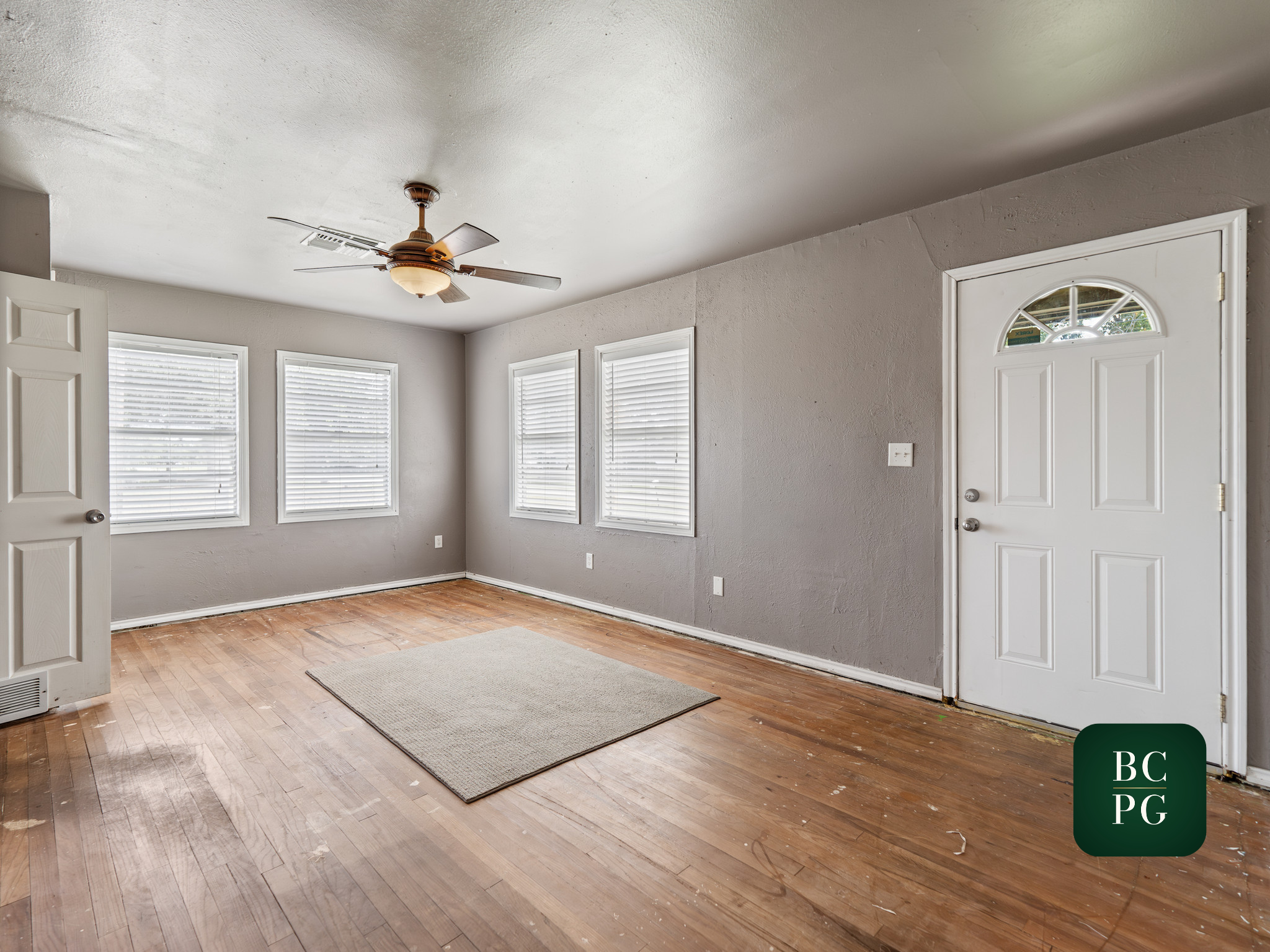 Empty living room with hardwood floors, light gray walls, white window blinds, a ceiling fan, and a front door with a semi-circular window.