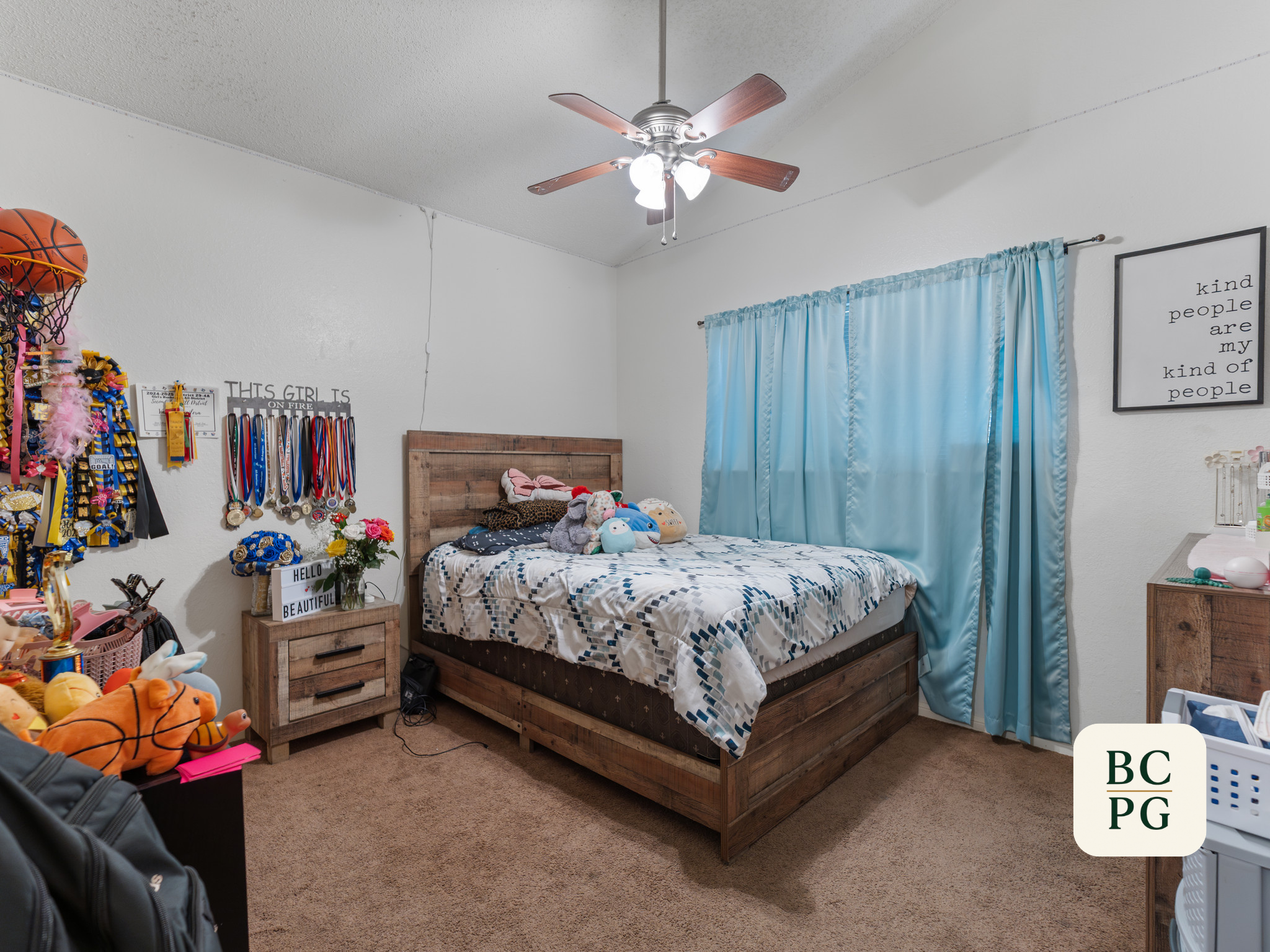 A bedroom featuring a wooden bed with stuffed animals, a blue curtain, and a white wall with inspirational quote signs, along with various toys and medals on display.