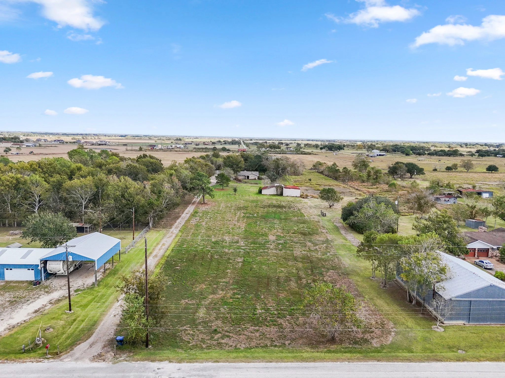 A rural landscape with open fields, scattered trees, and a few buildings under a blue sky with clouds.
