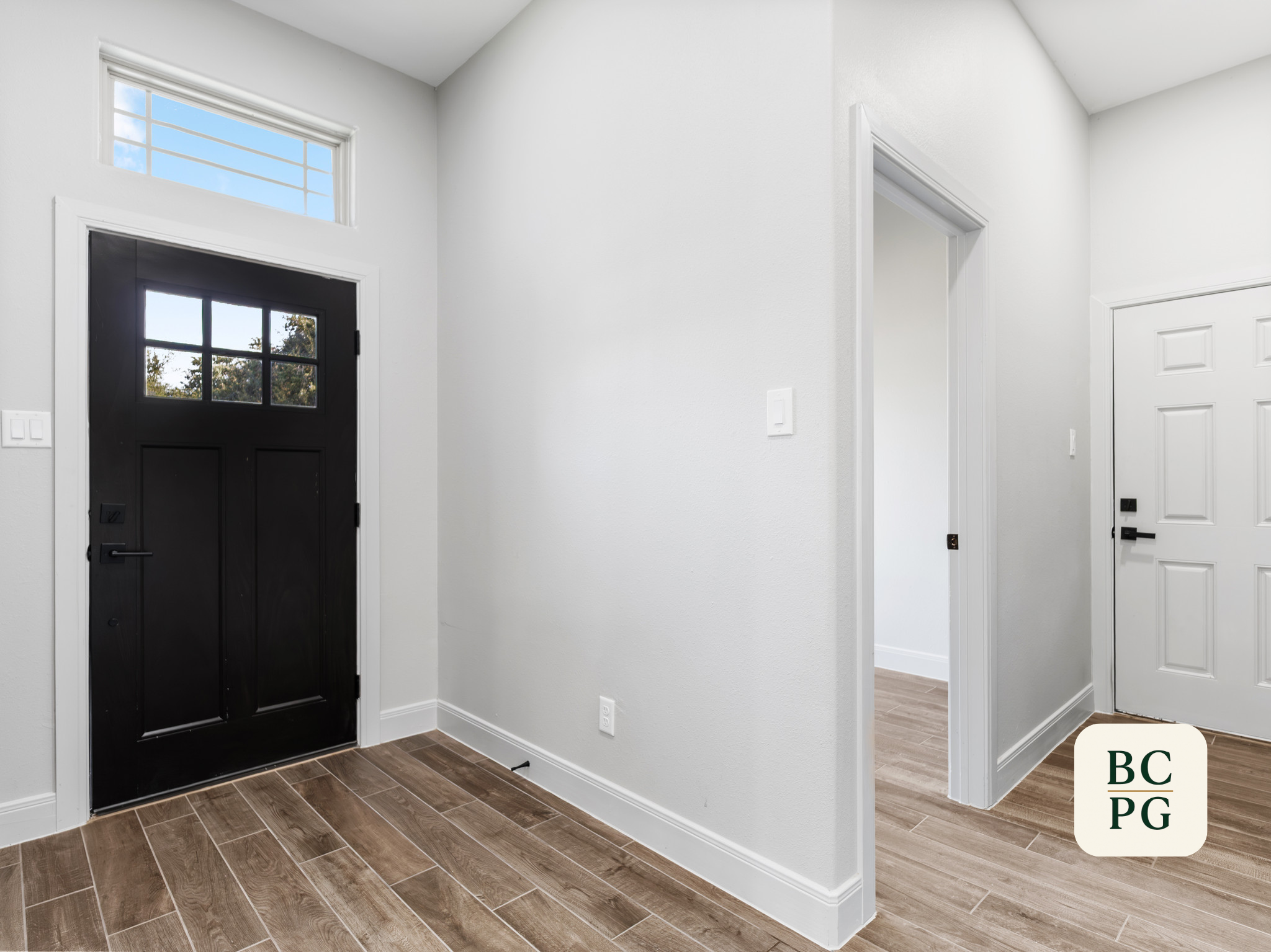 Interior of a modern home with a black front door and a white interior door, white walls, wooden flooring, and a transom window above the front door.