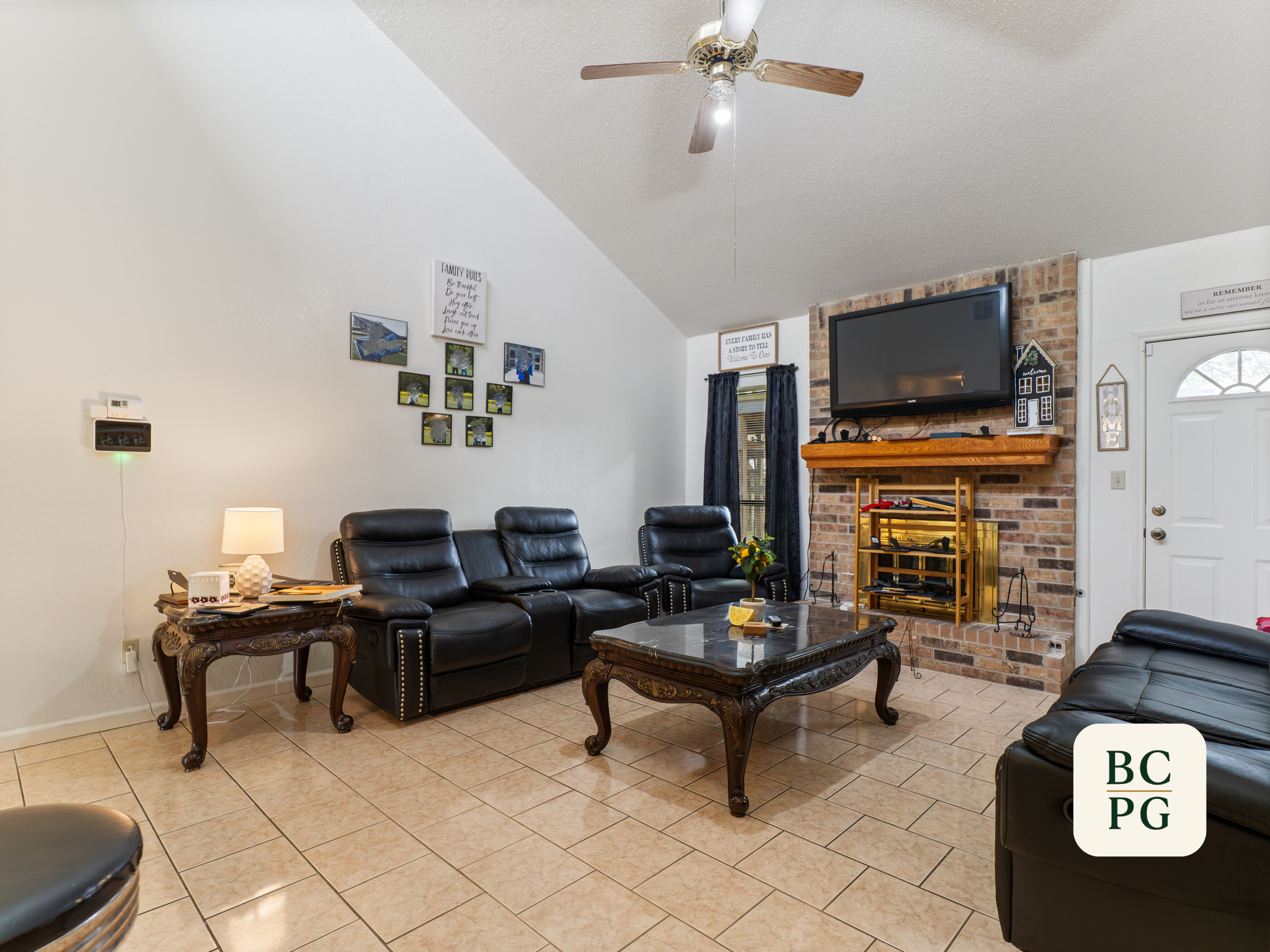 Living room with black leather sofas, a coffee table, a brick fireplace with a flat-screen TV mounted above it, and walls decorated with framed photos and signs.