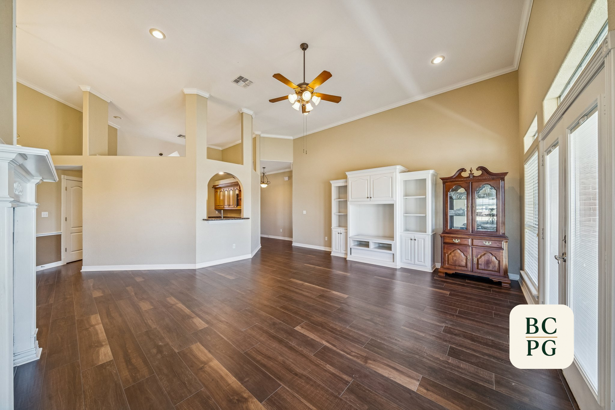 Interior view of a spacious living room with hardwood floors, white and beige walls, a ceiling fan with lights, large windows with blinds, and white built-in cabinets. There is a wooden china cabinet on the right side.