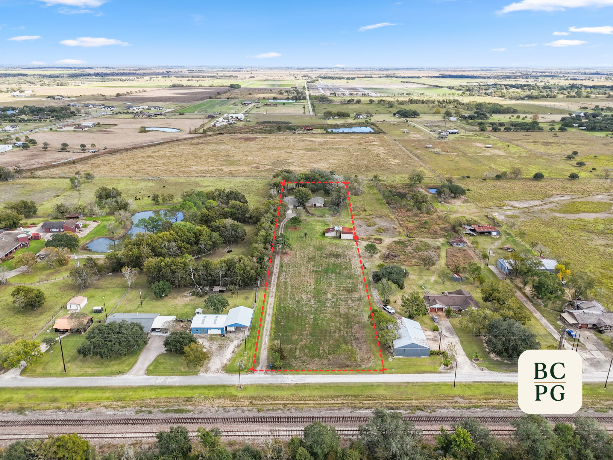 Aerial view of rural farmland with various houses, ponds, and fields. A property outlined in red highlights a specific parcel of land between other properties and a railway track. The surrounding landscape features flat terrain with scattered trees a