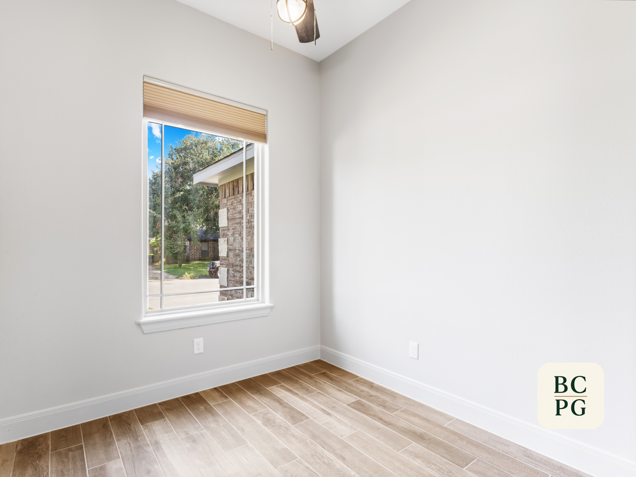 Empty room with white walls, a window with beige blinds showing a view of a house and trees outside, brown wood flooring, ceiling fan with light fixture.