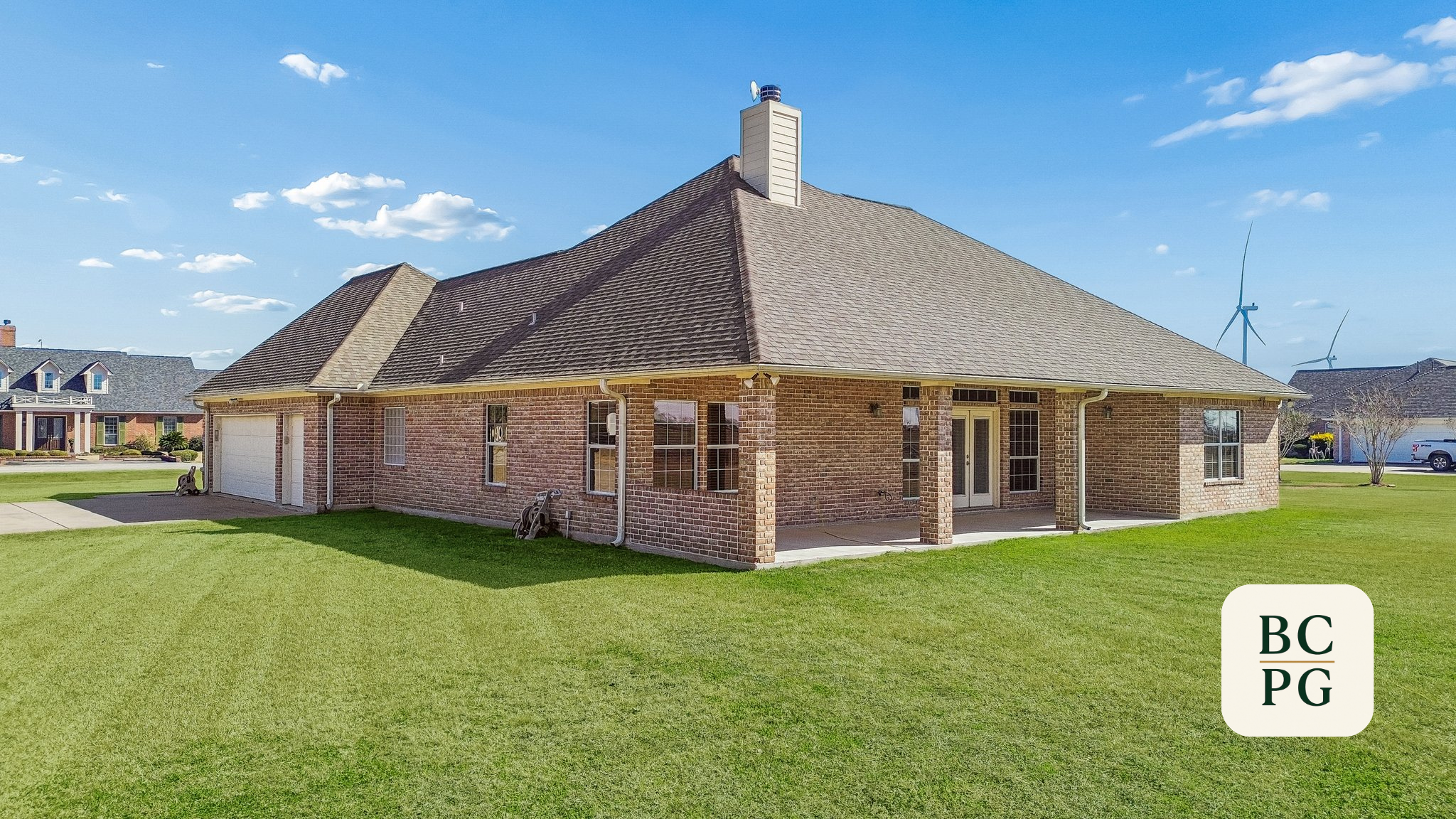 A brick house with a pitched roof, a chimney, multiple windows, a white garage door, and a front door, situated on a green lawn with neighboring houses in the background, under a blue sky with some clouds.