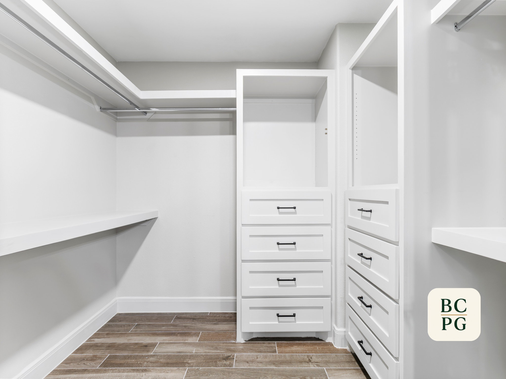 Empty walk-in closet with white shelves, drawers, and hanging rods, wood flooring, and white walls.