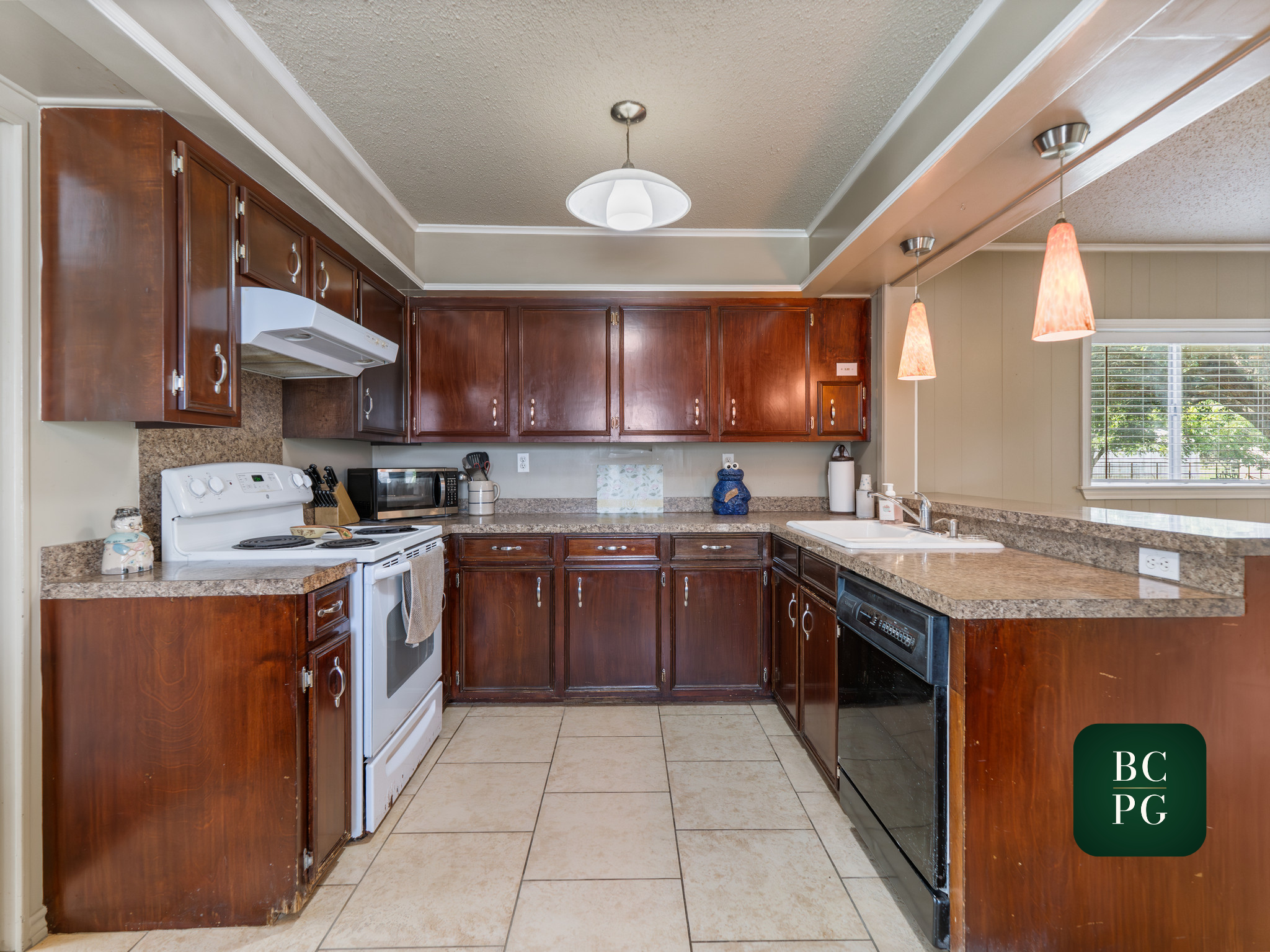 A kitchen with dark wooden cabinets, white appliances, granite countertops, beige tiled floor, and a window with blinds. There are hanging pendant lights and a ceiling light fixture.
