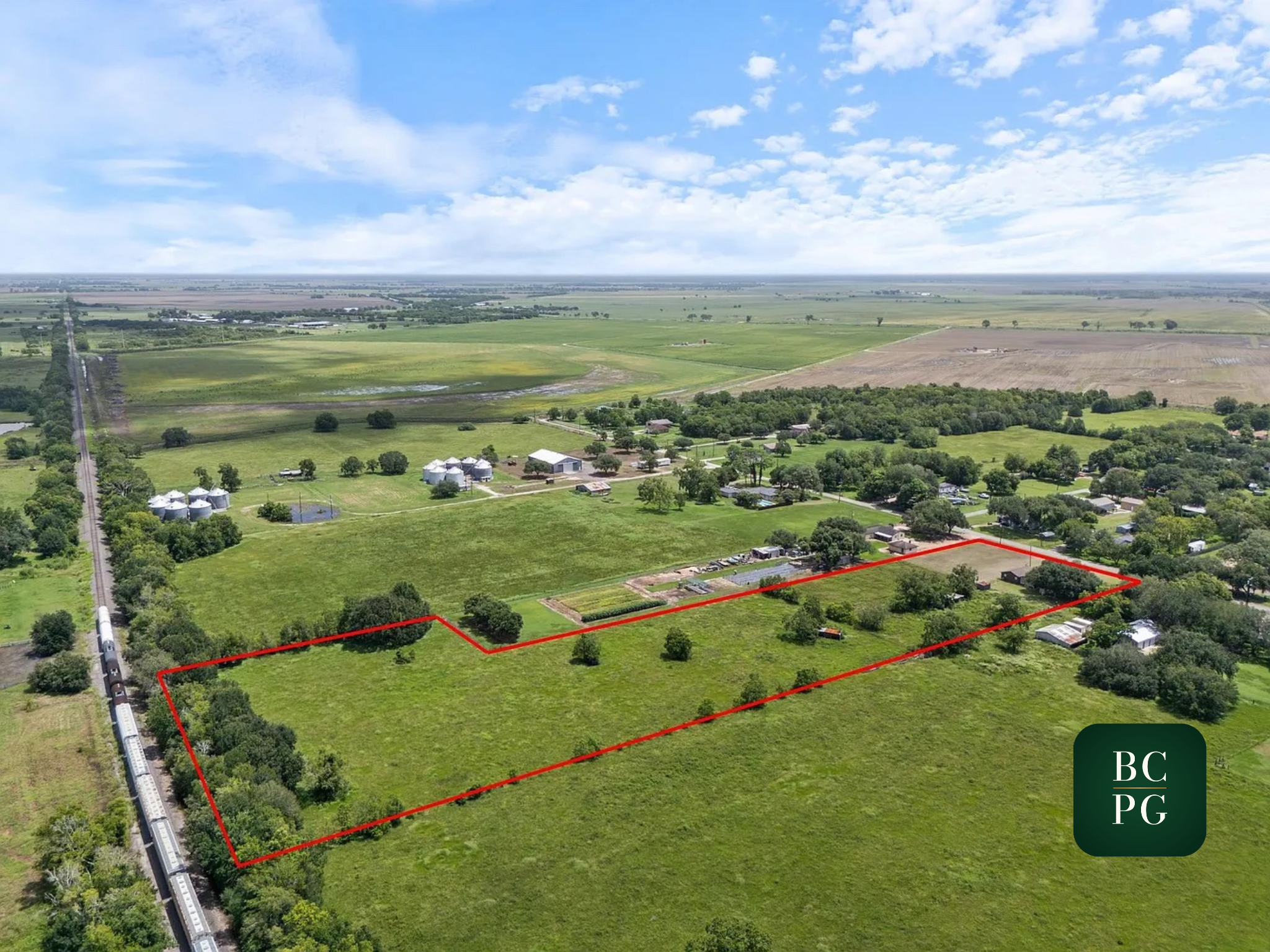 Aerial view of green farmland with properties, trees, and a train on the left rail line, under a blue sky with scattered clouds, with a section outlined in red.