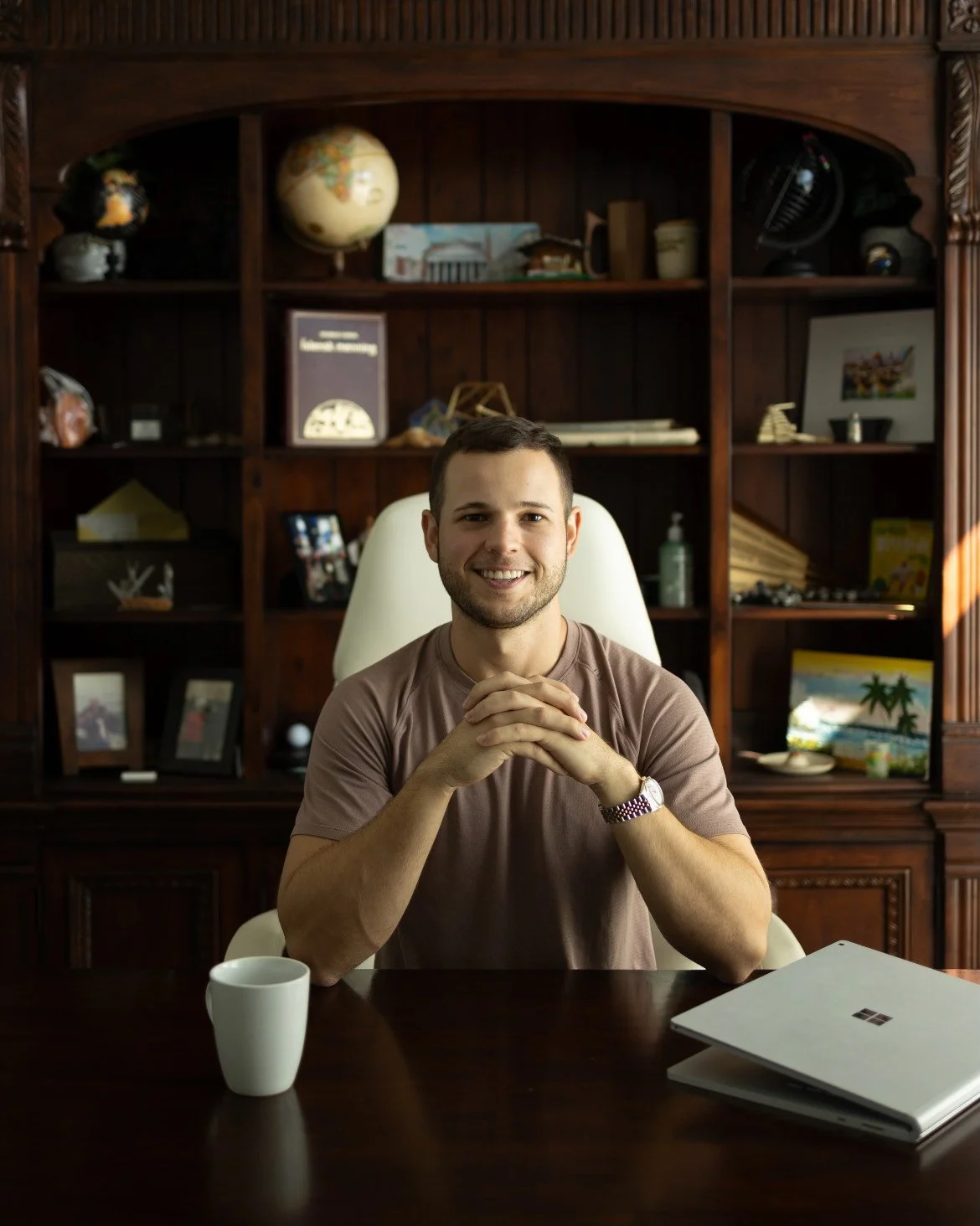 A man sitting at a desk, smiling with hands clasped, in a room with a large wooden bookshelf behind him filled with various objects and framed photographs.