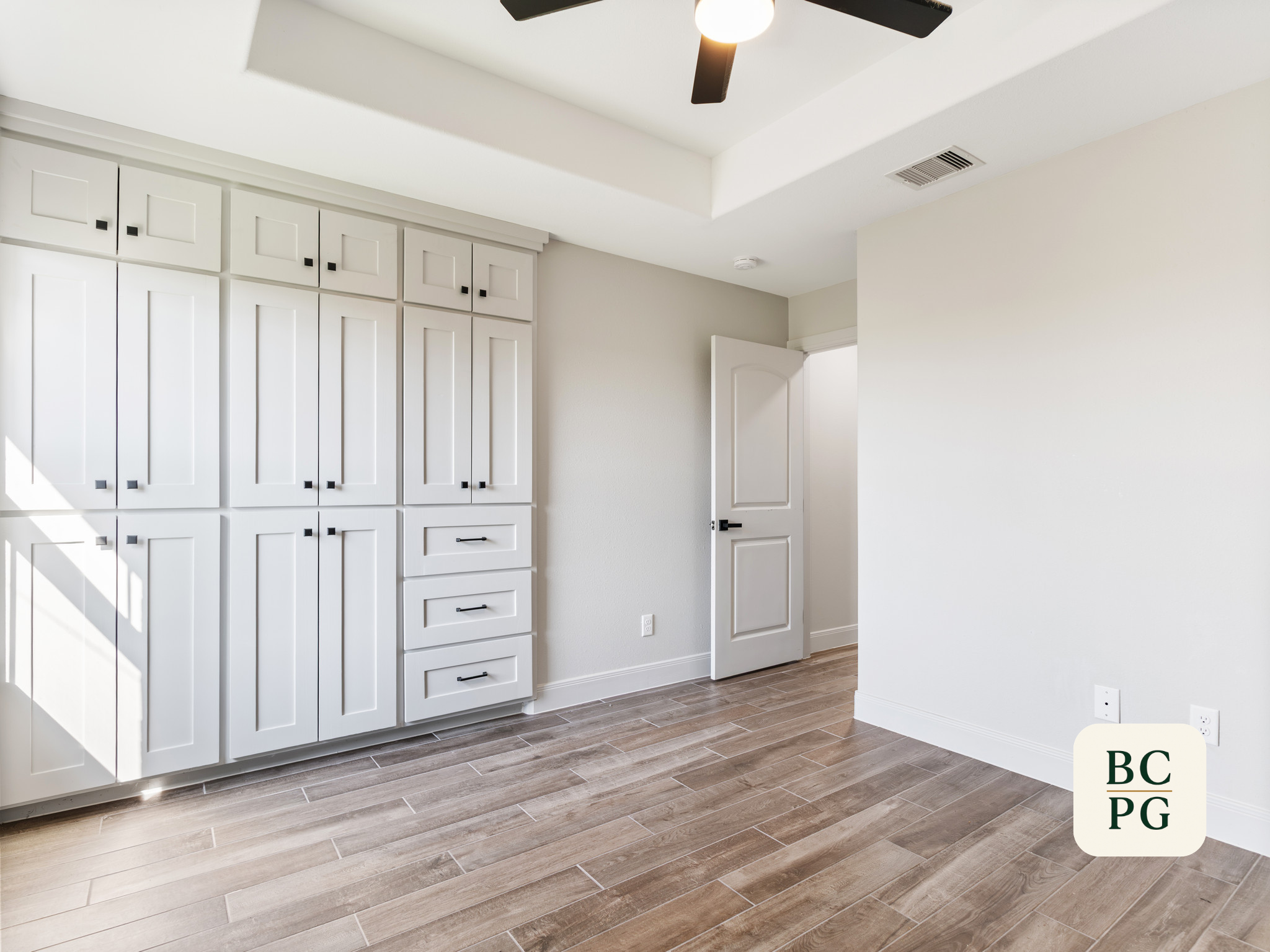 A room with a built-in white cabinet with multiple doors and drawers, a white open door, a ceiling fan with black blades, a ceiling vent, and wood-like floor tiles.