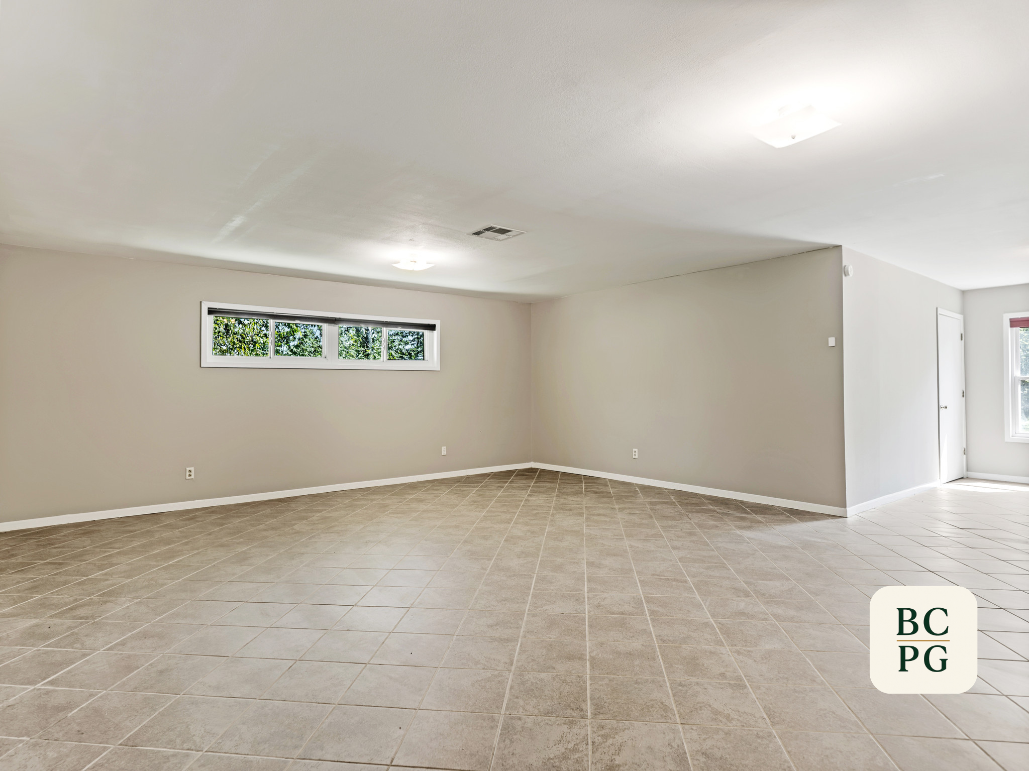 Empty living room with beige tiled floor, light-colored walls, rectangular window with green foliage outside, ceiling lights, and vent.
