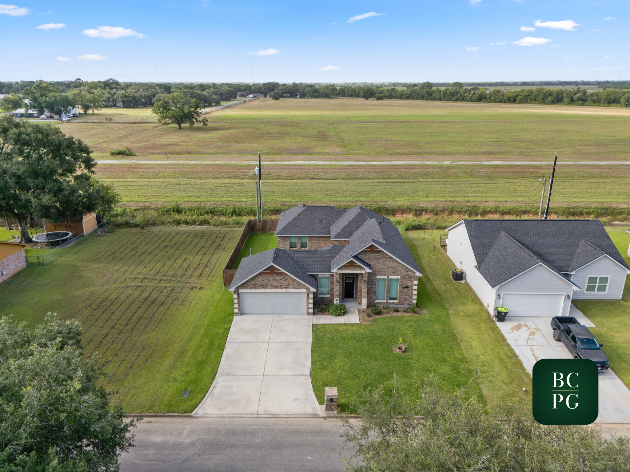 A suburban house with a well-maintained front yard, concrete driveway, and neighboring houses, with fields and trees in the background under a blue sky with clouds.