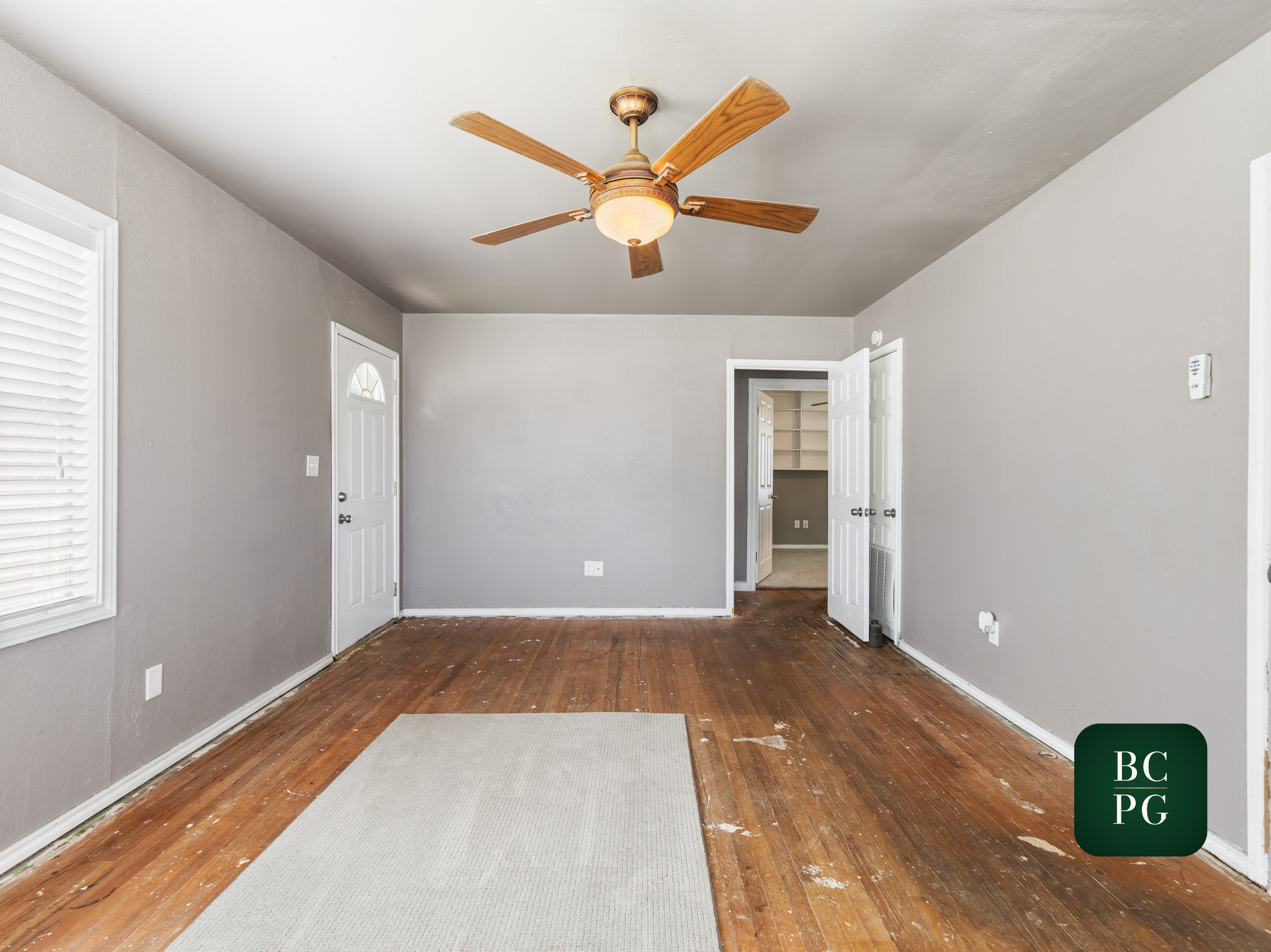 Empty living room with hardwood floors, light gray walls, white window blinds, and an open double door leading to another room with built-in shelves.