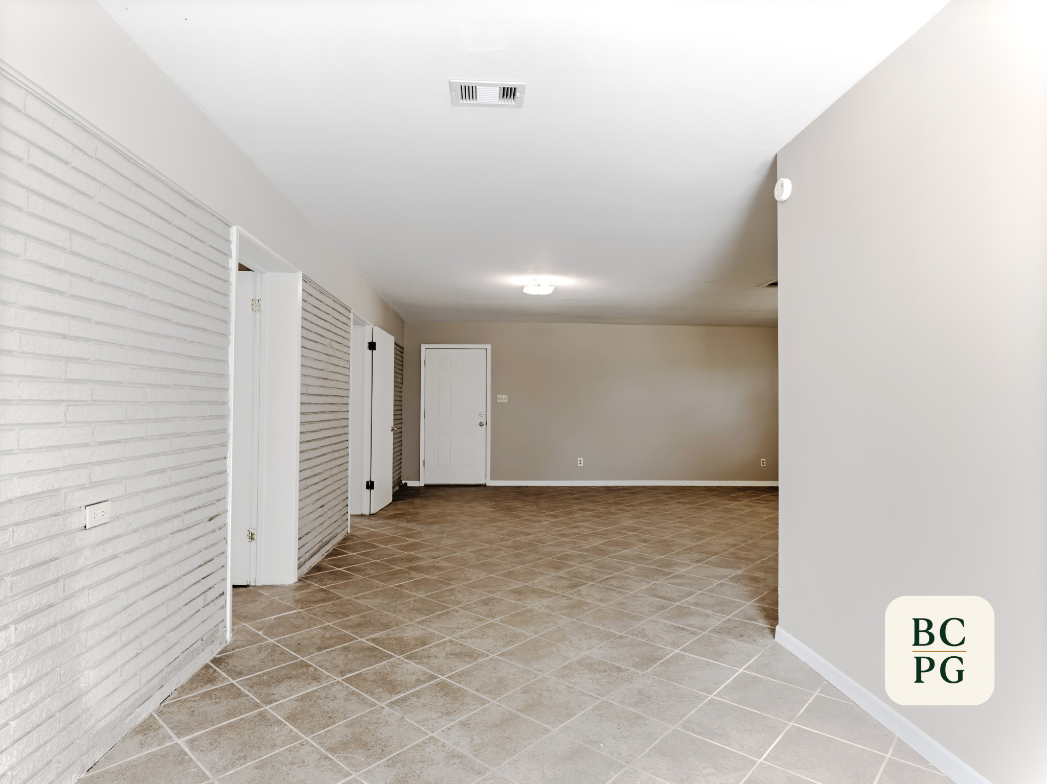 Interior view of a room with light-colored walls, a white brick accent wall, a white ceiling with a vent and a ceiling light, brown tile flooring, and doors on the left side.