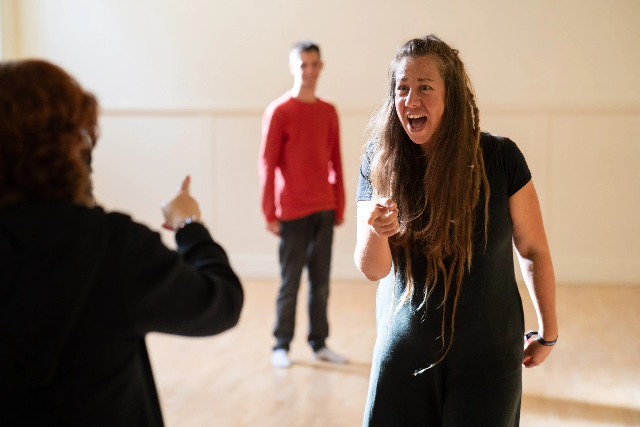 Lois is in the studio interacting with a participant. she is wearing all black, in the background another participant is onlooking wearing a red top and black trousers.