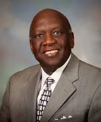 A portrait of a middle-aged African American man with gray hair and beard, dressed in a dark suit, white shirt, and patterned tie, against a mottled gray background.