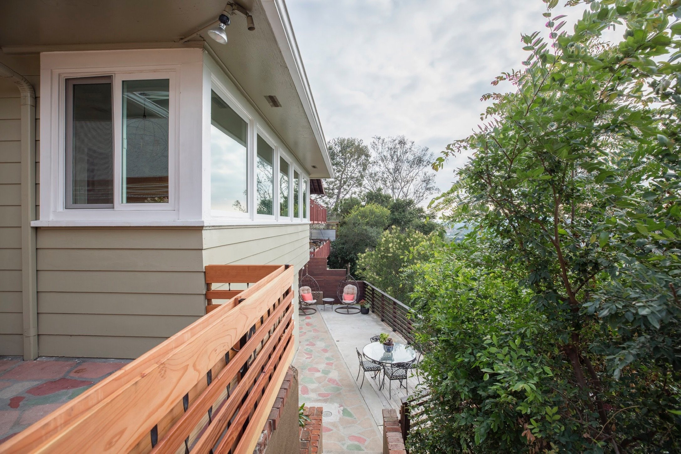 Exterior of a house with large windows and a patio. The patio includes a metal table with chairs and two hanging chairs. Lush greenery surrounds the area.