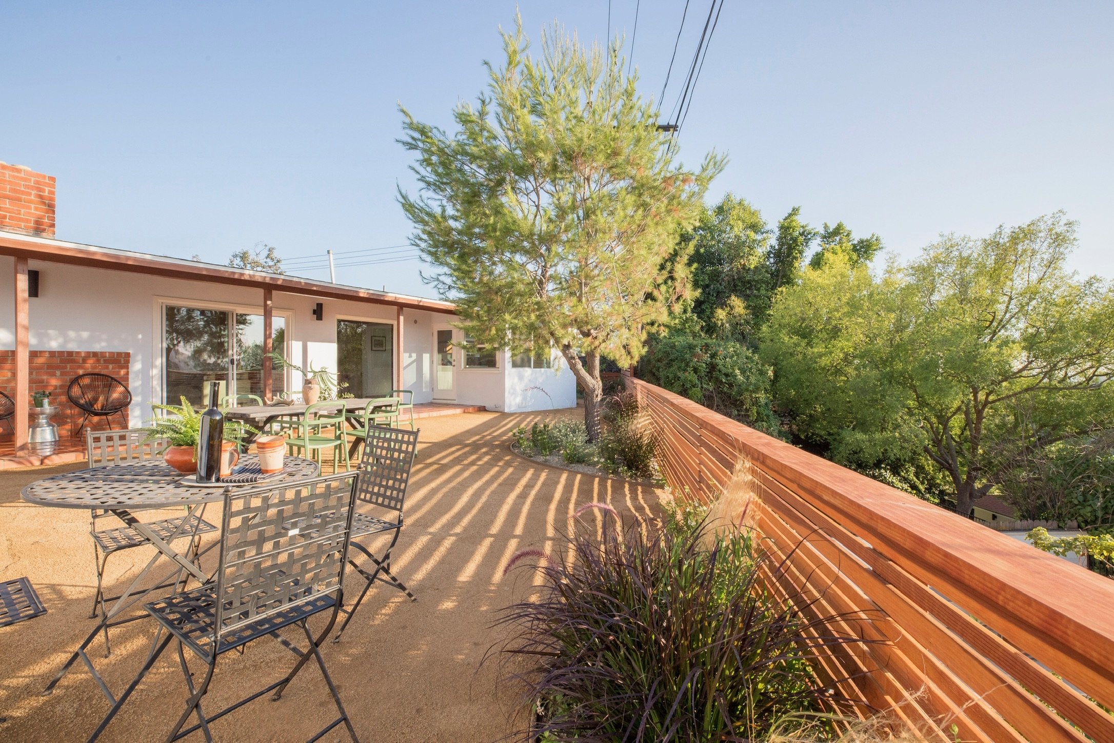 Outdoor patio with metal tables and chairs, potted plants on tables, surrounded by wooden fence and trees, adjacent to a building with large windows.
