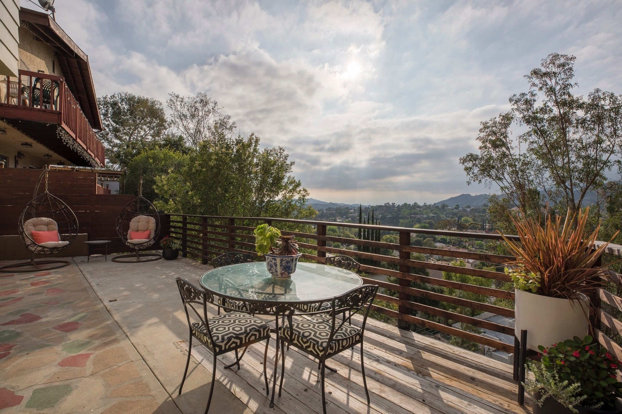 Outdoor patio with glass table and chairs, hanging egg chairs, potted plants, wooden railing, and scenic view of trees and hills under a cloudy sky.
