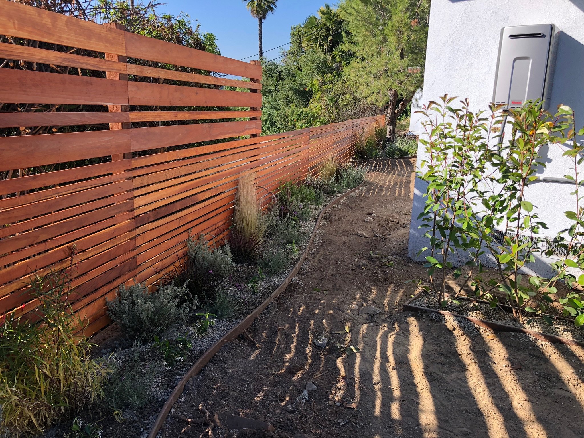 Outdoor area with a wooden slat fence, dirt path, and various plants alongside a white building.