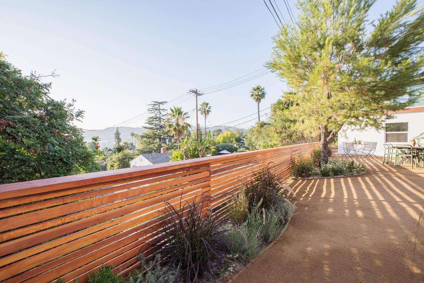 Outdoor patio area with wooden slat fence, trees, and plants. Patio furniture includes tables and chairs. Background features palm trees and distant hills under a clear sky.