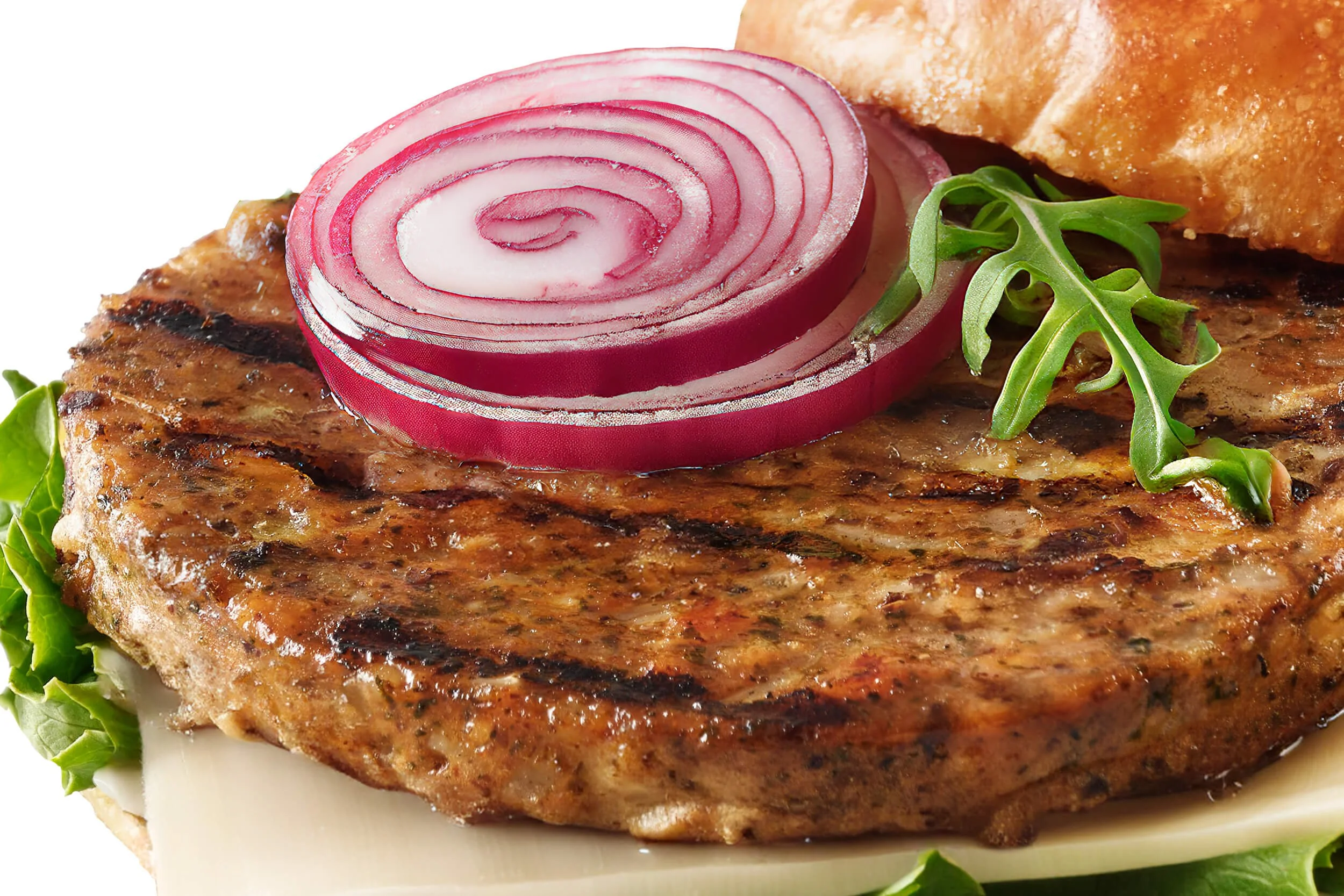 Close-up of a grilled hamburger patty topped with red onion slices and arugula, with a bread bun in the background.