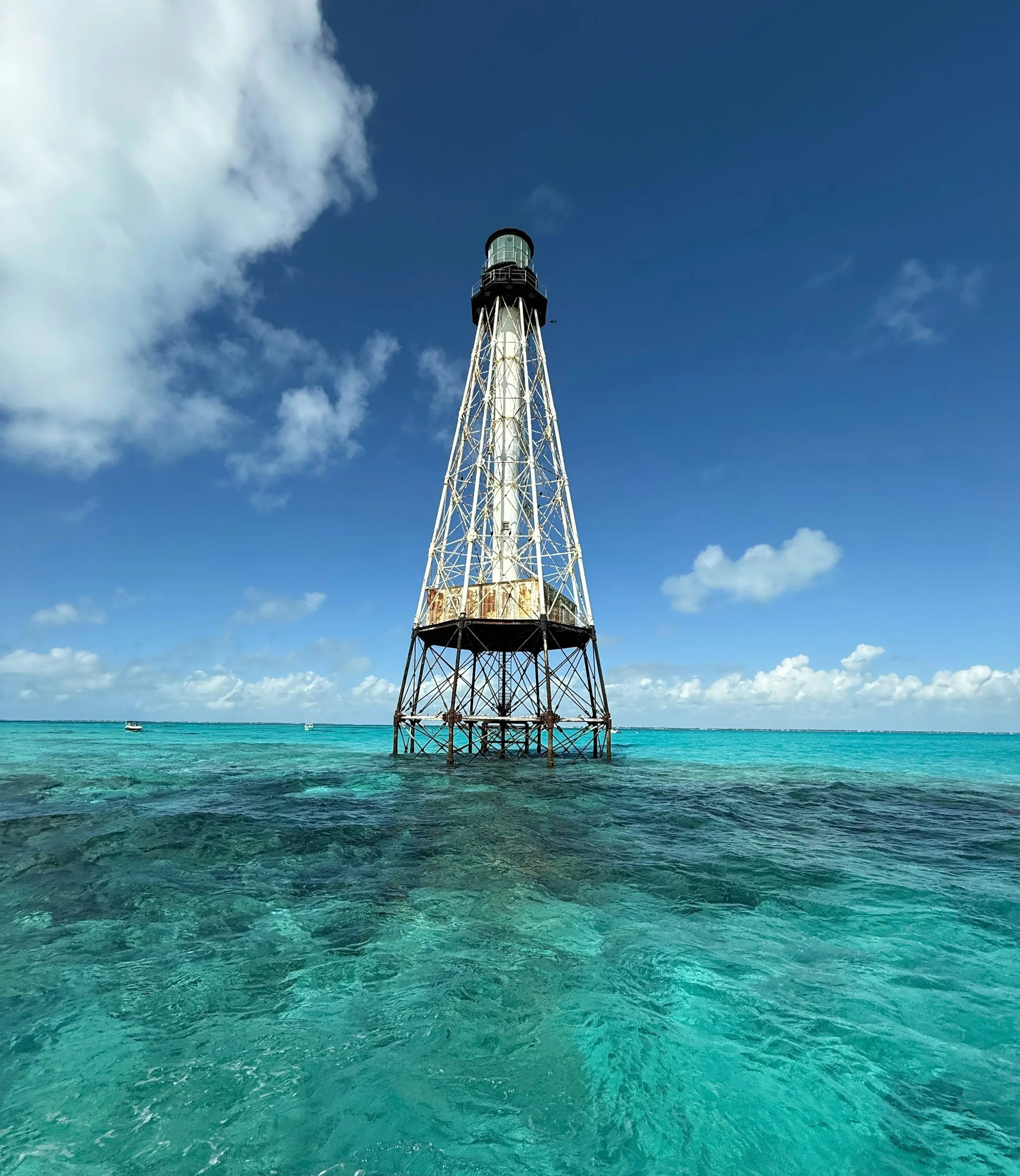 Tall lighthouse structure in the ocean with clear blue sky and white clouds in the background.