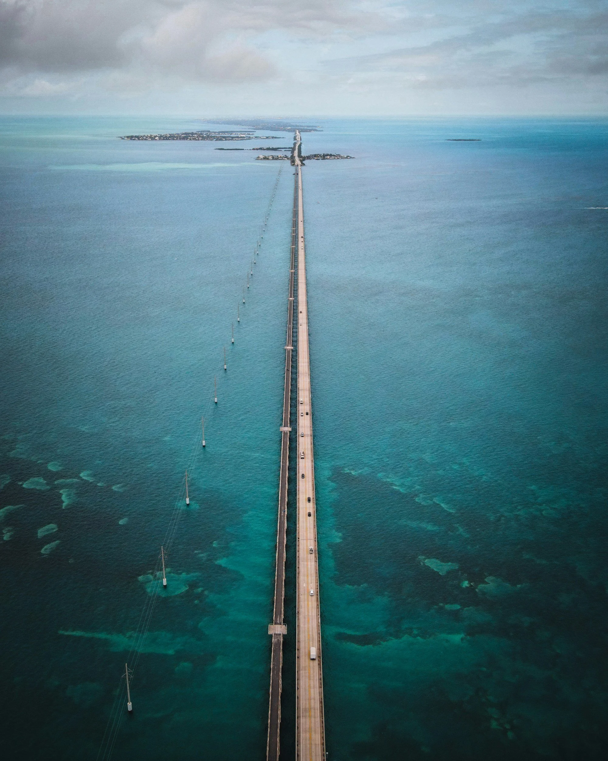 An aerial view of a long bridge extending over turquoise ocean waters, with small islands and a town visible in the distance under cloudy sky.