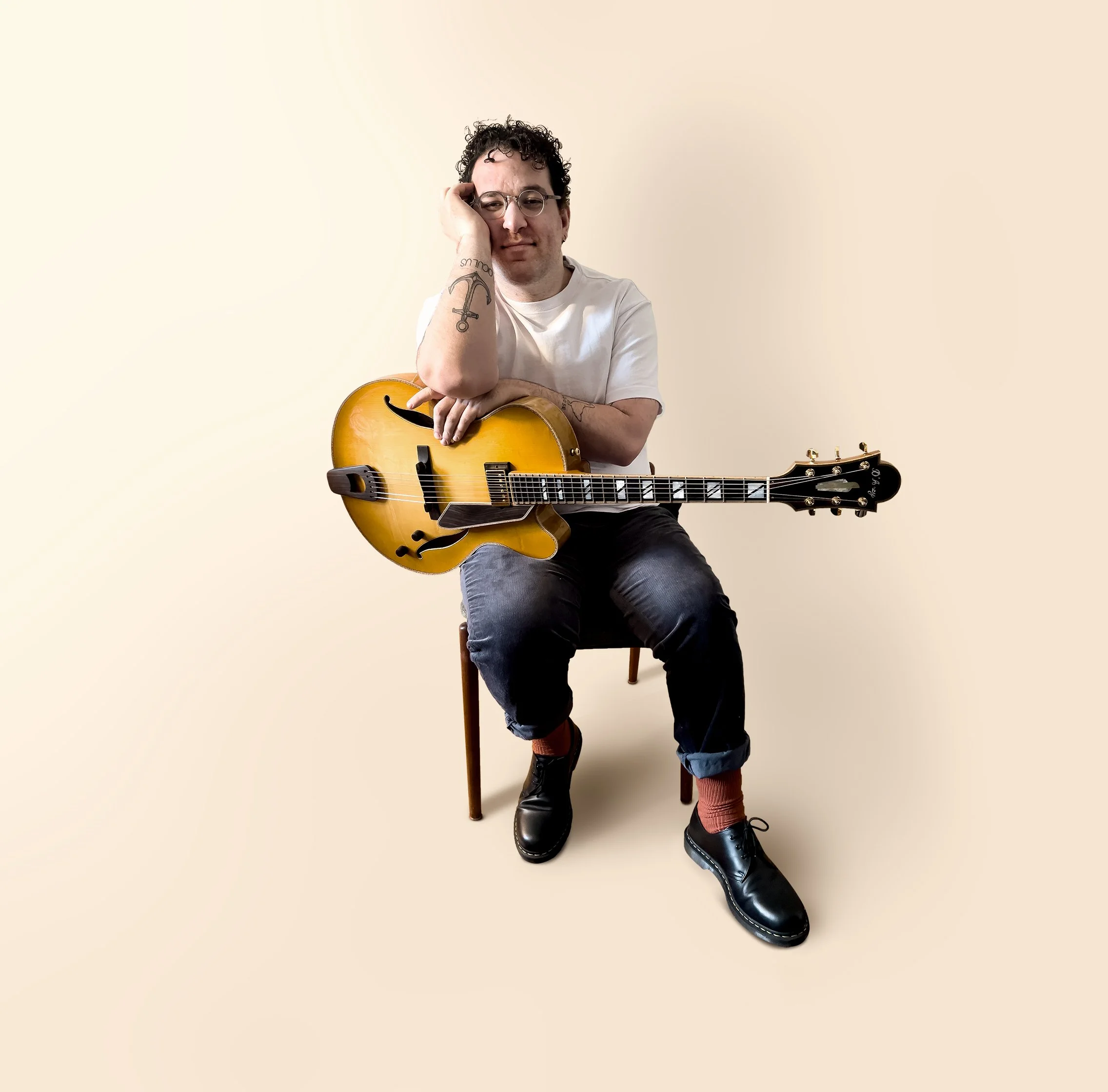 Person sitting on a chair holding a yellow electric guitar, wearing glasses, a white t-shirt, jeans, and black shoes with red socks, on a light beige background.