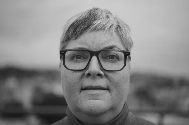 Black-and-white close-up photo of a woman with short hair and glasses, outdoors with a blurred landscape background.