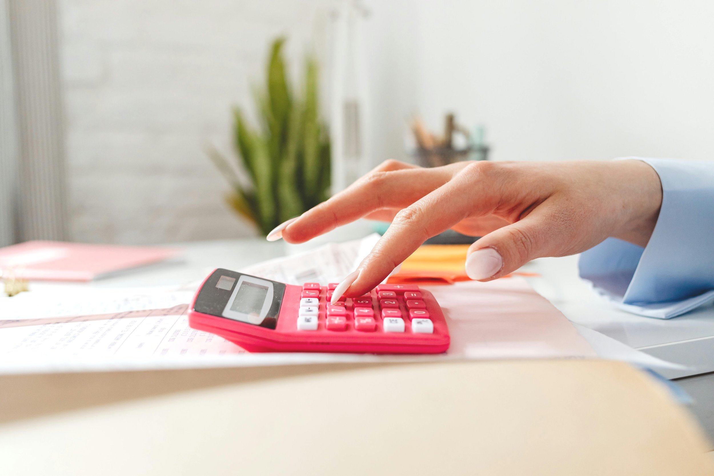 A person using a pink calculator on a cluttered desk with documents and a potted plant in the background.