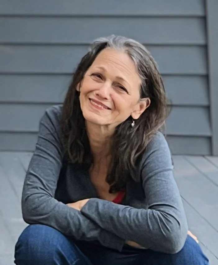 A woman with gray hair smiling and sitting outdoors in front of a gray wooden wall.