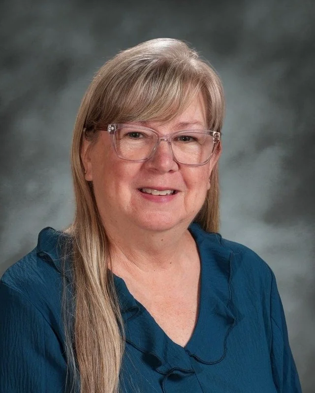 A school crest featuring a shield divided into four sections with a cross, a stylized crown, a woman with a halo, a diploma scroll, and a dove. Beneath the shield is a banner with the Latin phrase "Adora Disce Serve" and the year "1983." Below that, the text reads "Coming Soon."