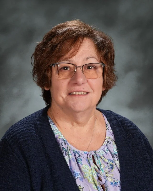 A crest with a cross, a crown, a woman with a halo, a rolled scroll, and a dove, with the words "Adora Disce Serve" and the year "1983". Below the crest, it says "Coming Soon."