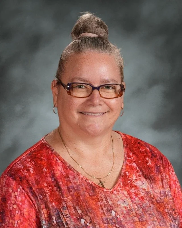 A digital graphic with a shield emblem divided into four sections, featuring a cross with a crown, a woman with a halo, a rolled document, and a dove. Below the shield is a banner with the Latin phrase "Adora Disce Serve" and the year 1983, followed by the words "Coming Soon."