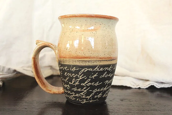 Claire Lemp Ceramics. Bozeman, MT Potter, pottery.  A ceramic mug with handwritten script and a beige glaze sitting on a dark surface with a white cloth in the background.