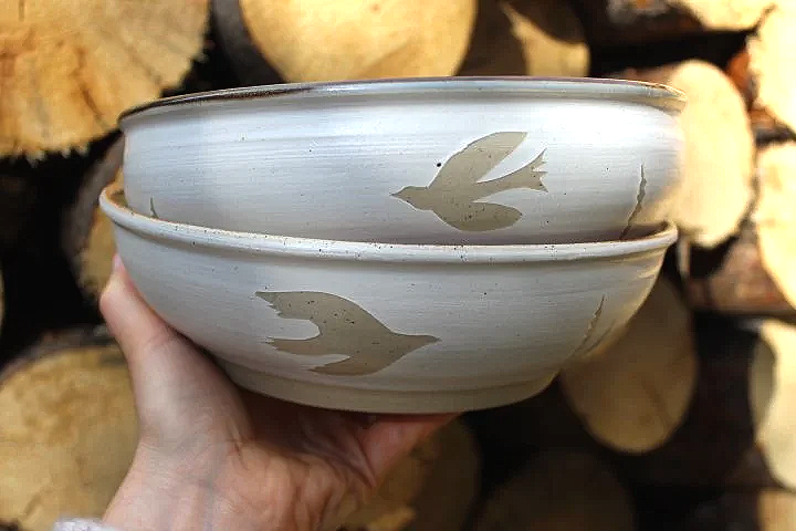 Claire Lemp Ceramics. Bozeman, MT Potter, pottery.  Two ceramic bowls with bird designs being held in a person's hand, with stacked firewood in the background.