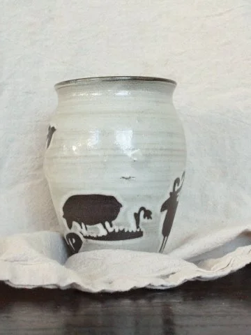 Claire Lemp Ceramics. Bozeman, MT Potter, pottery.  A ceramic vase with black animal and plant designs, placed on a white cloth on a wooden surface.