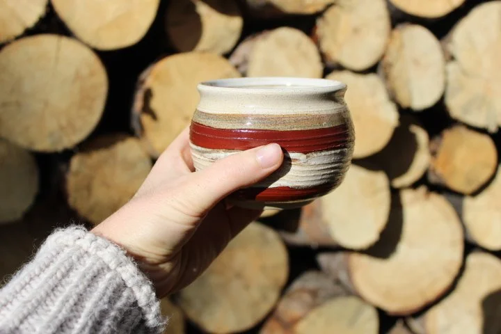 Claire Lemp Ceramics. Bozeman, MT Potter, pottery. Montana Ceramics, ceramicist. Made In Montana. A person holding a ceramic bowl with red and beige stripes in front of a stacked woodpile.