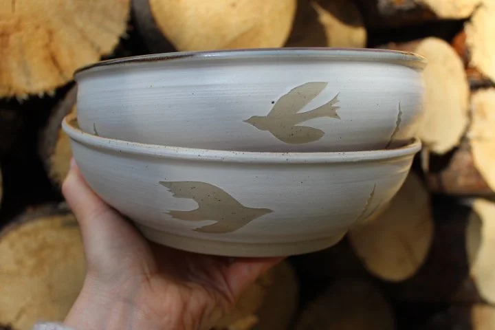 Claire Lemp Ceramics. Bozeman, MT Potter, pottery.  A hand holding two stacked ceramic bowls with a bird silhouette design, with a background of stacked logs.