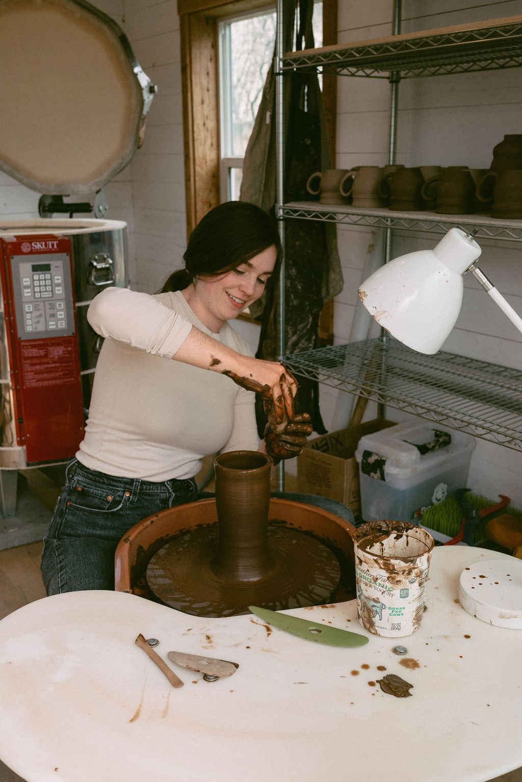 Claire Lemp Ceramics. Bozeman, MT Potter, pottery. A woman in a white shirt and jeans is shaping a tall pottery piece on a potter's wheel, with chocolate glaze covering her hands and the pottery.