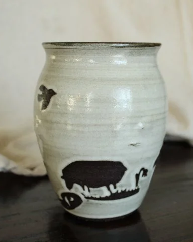 Claire Lemp Ceramics. Shepherd Mug. A ceramic vase with a white glaze and black animal silhouette designs, including a pig and other farm animals, sitting on a dark surface with a light background.