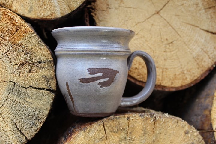 Claire Lemp Ceramics. Bozeman, MT Potter, pottery. Montana Ceramics, ceramicist. Made In Montana. Gray ceramic mug with a decorative handle resting against cut logs of wood in the background.