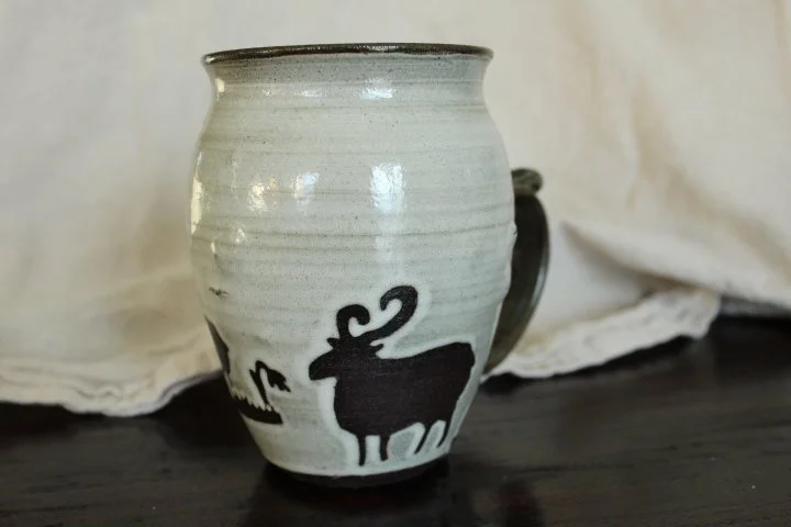 Claire Lemp Ceramics. Bozeman, MT Potter, pottery.  A ceramic mug with a black silhouette of a goat on a white glaze background, placed on a dark surface with white fabric in the background.