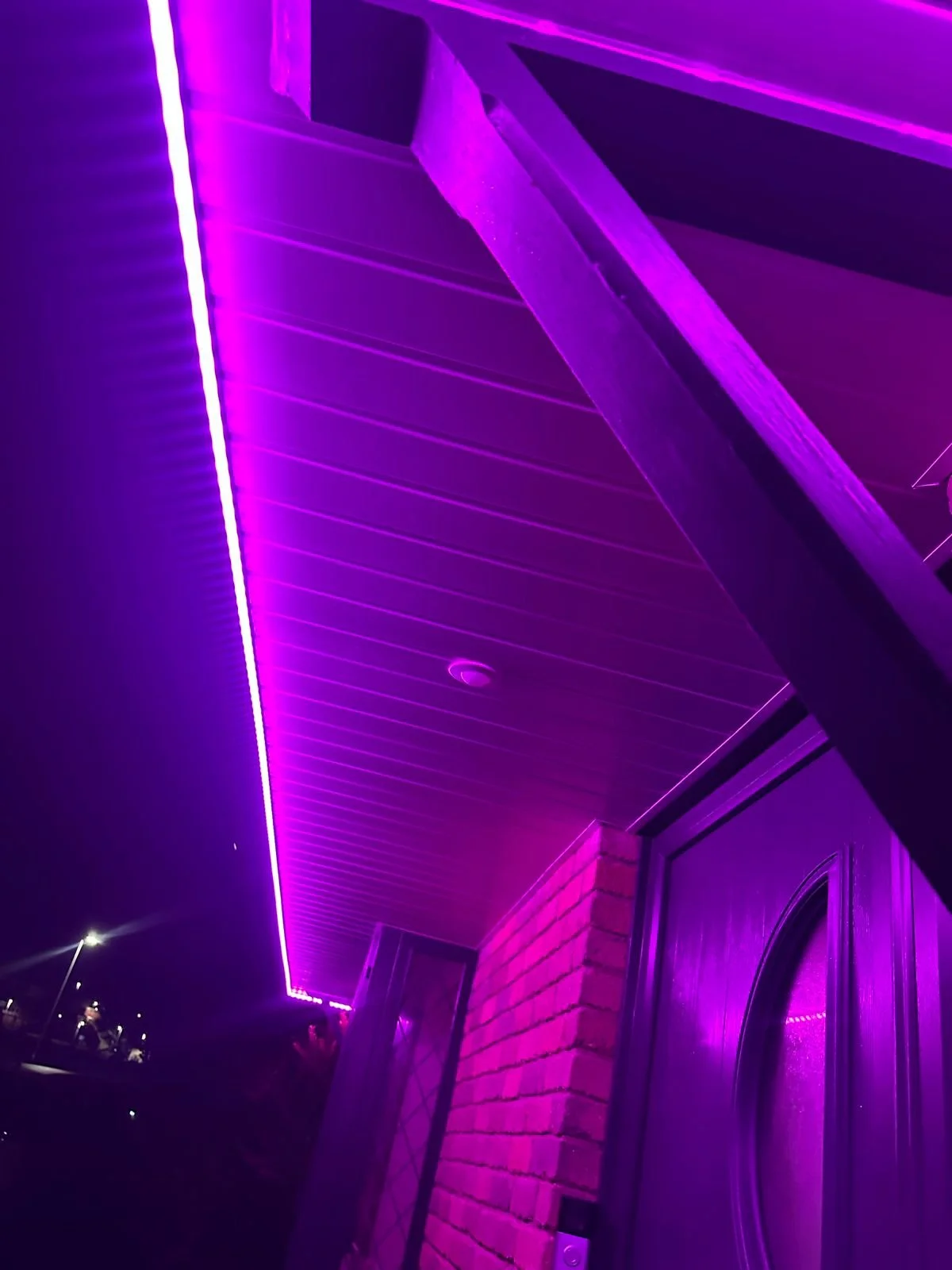 Close-up of a house exterior at night illuminated with vibrant purple LED strip lighting along the roofline, brick wall, and small part of a door.