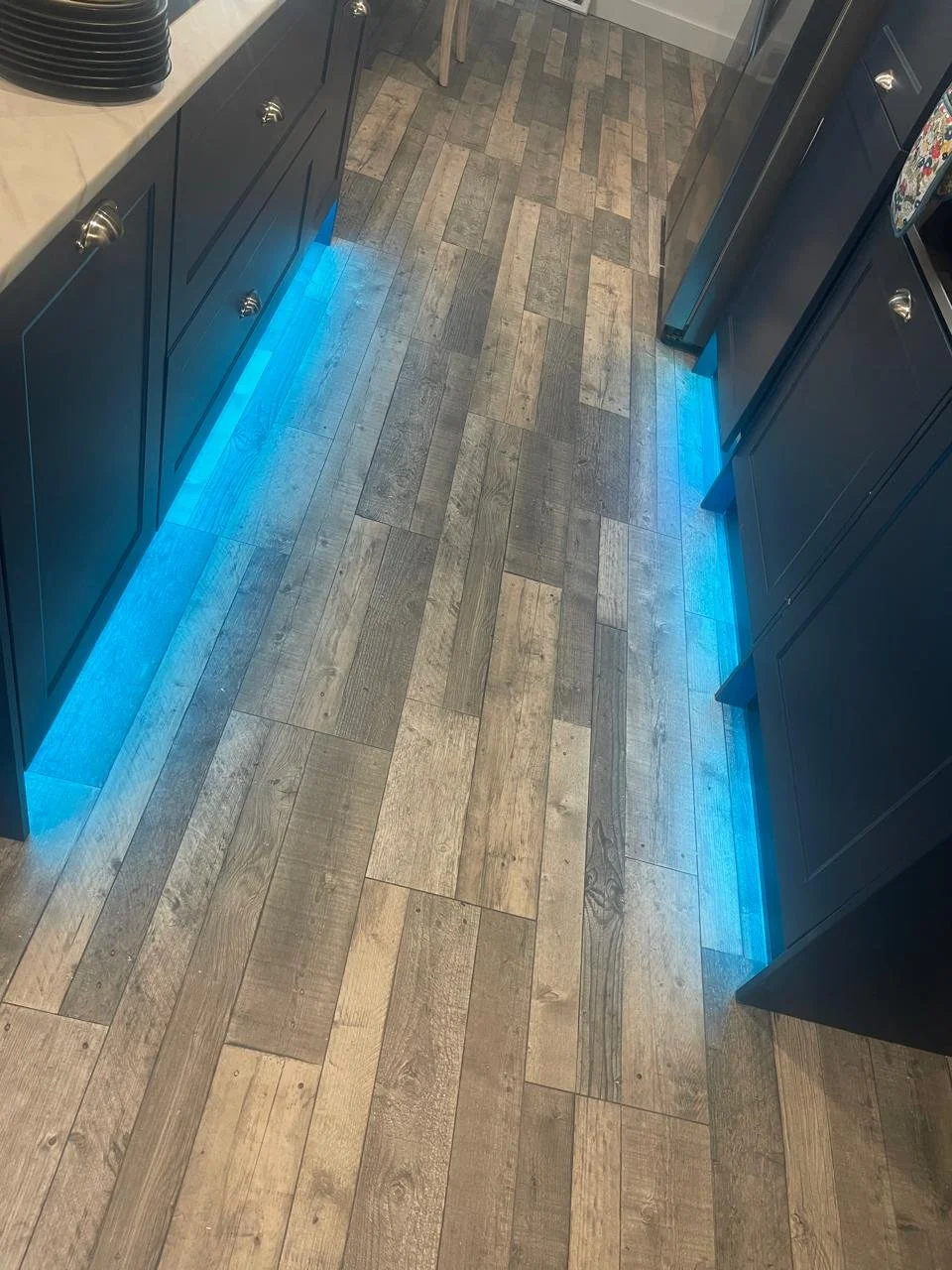 Kitchen floor with wooden planks and blue LED lights underneath the dark cabinets on both sides.